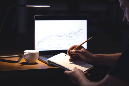 Un hombre escribe en un cuaderno con un lápiz frente a una computadora portátil que muestra un gráfico de líneas ascendente.