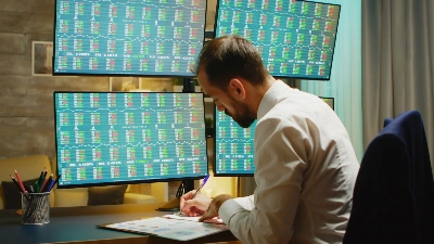 hombre concentrado en su escritorio de trabajo, rodeado de múltiples monitores que exhiben complejos gráficos de mercados financieros en tiempo real.