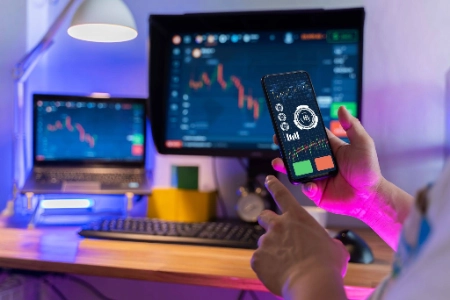 estación de trabajo dedicada al trading, con un enfoque en una persona sosteniendo un smartphone que muestra gráficos financieros y botones de operación. Al fondo, se aprecian un monitor principal y una laptop, ambos exhibiendo plataformas de análisis con velas japonesas en un entorno iluminado por luces neón de color púrpura y azu