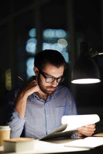 hombre joven con barba y gafas, profundamente concentrado mientras revisa documentos físicos bajo la luz de una lámpara de escritorio.