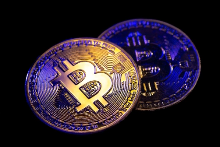 Dos monedas físicas de Bitcoin aparecen sobre un fondo negro profundo, iluminadas con luces de color azul y dorado.