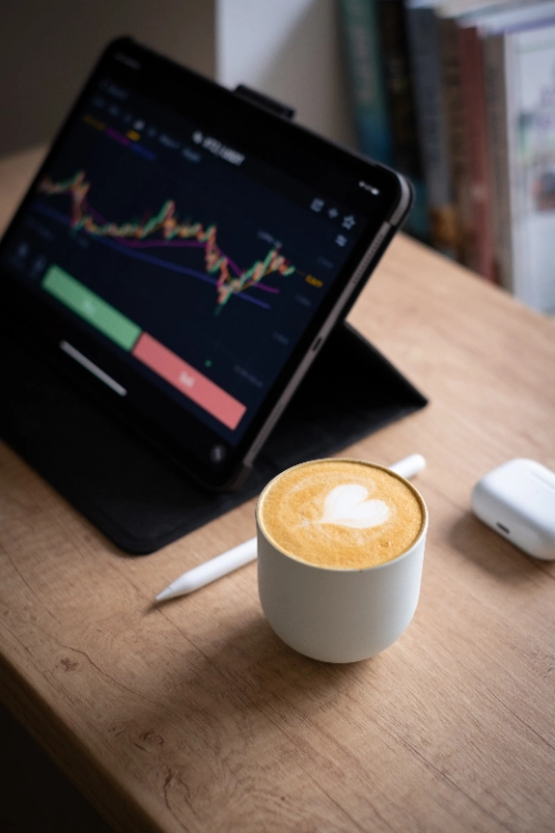 Una taza de café con arte latte de corazón en primer plano sobre una mesa de madera, junto a un lápiz digital, unos audífonos inalámbricos y una tableta que muestra gráficos financieros de 'trading'.