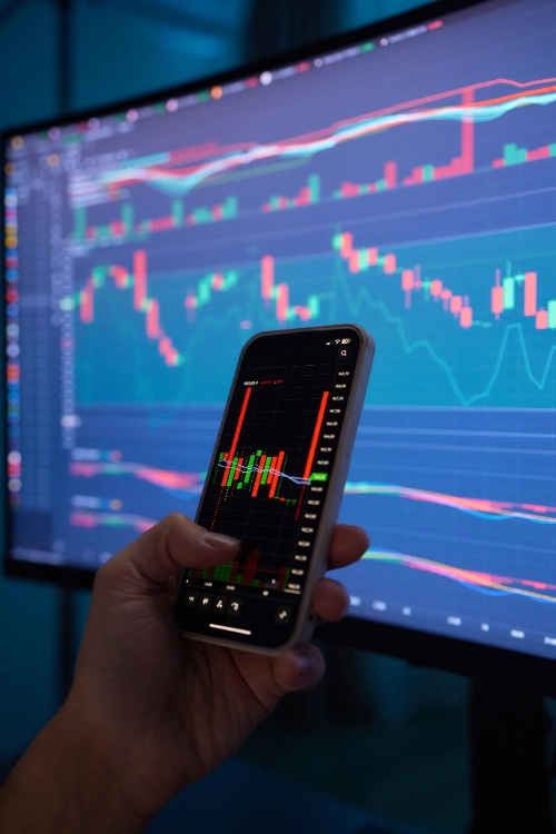 se muestra en primer plano una mano sosteniendo un teléfono celular con un gráfico de velas financieras, frente a un gran monitor que despliega pantallas de trading adicionales bajo una iluminación azul de fondo.