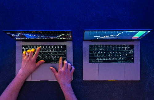 Vista cenital de las manos de una persona operando simultáneamente en dos computadoras portátiles abiertas que muestran gráficos financieros de trading