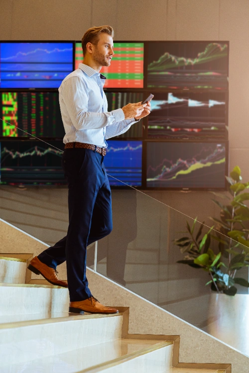 hombre con camisa clara y pantalón oscuro camina por unas escaleras mientras sostiene un teléfono móvil, con grandes pantallas de gráficos financieros de fondo.