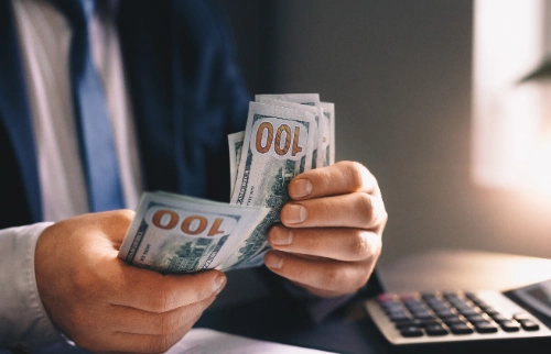 Un hombre con traje cuenta un mazo de billetes de cien dólares frente a una calculadora sobre su escritorio.