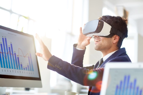 Un hombre con traje azul usa un visor de realidad virtual frente a una computadora. En las pantallas de fondo se muestran gráficos de barras azules.