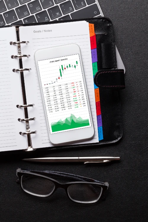 Vista cenital de un teléfono móvil con gráficos de análisis bursátil sobre una agenda abierta, acompañados por un bolígrafo, gafas de lectura y parte de un teclado en un escritorio oscuro.