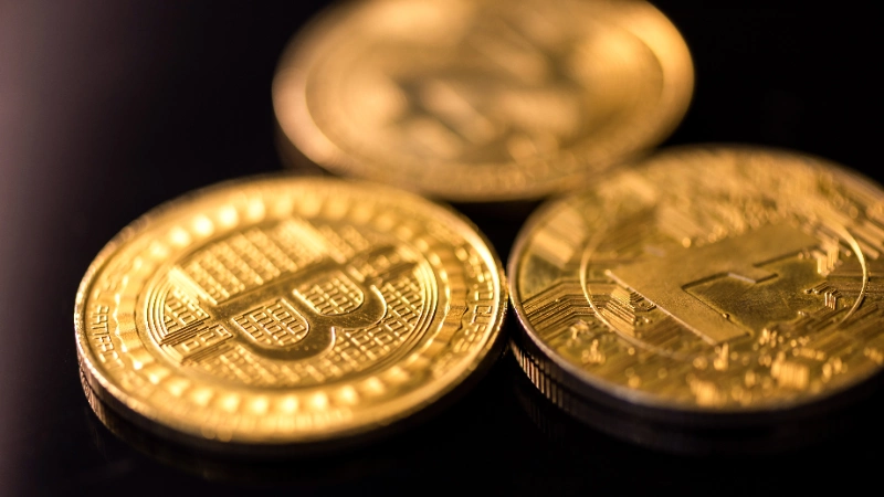 Tres monedas doradas de criptomonedas, entre ellas Bitcoin y Litecoin, reposan agrupadas sobre una superficie oscura y reflectante.