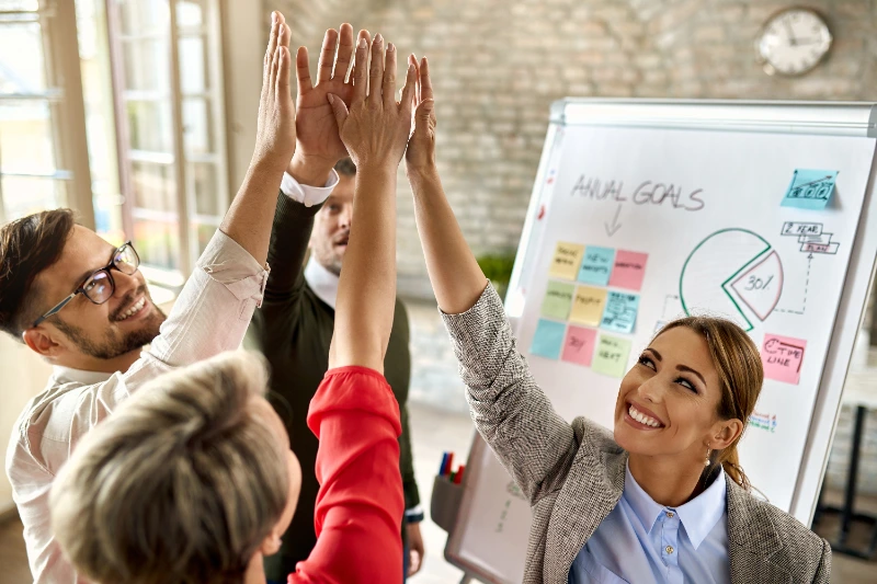 Un equipo de profesionales sonrientes celebra sus logros juntando sus manos en un chócala (high-five) frente a una pizarra con metas anuales y diagramas.