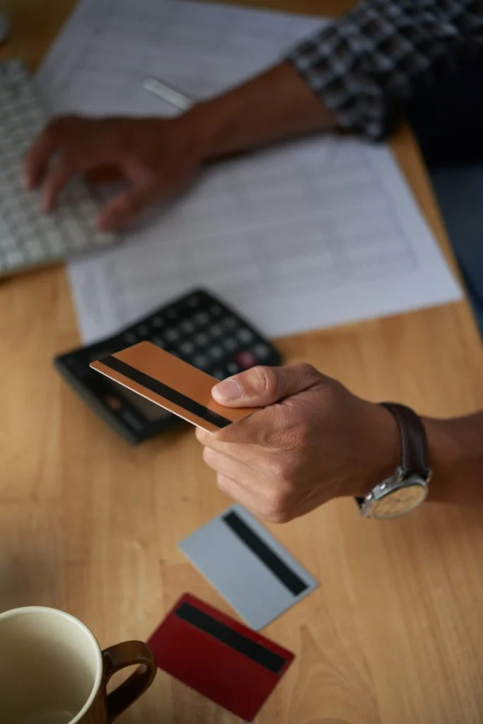 Un primer plano en ángulo picado de una persona sosteniendo una tarjeta de crédito dorada frente a un teclado, una calculadora y otras tarjetas sobre un escritorio de madera.