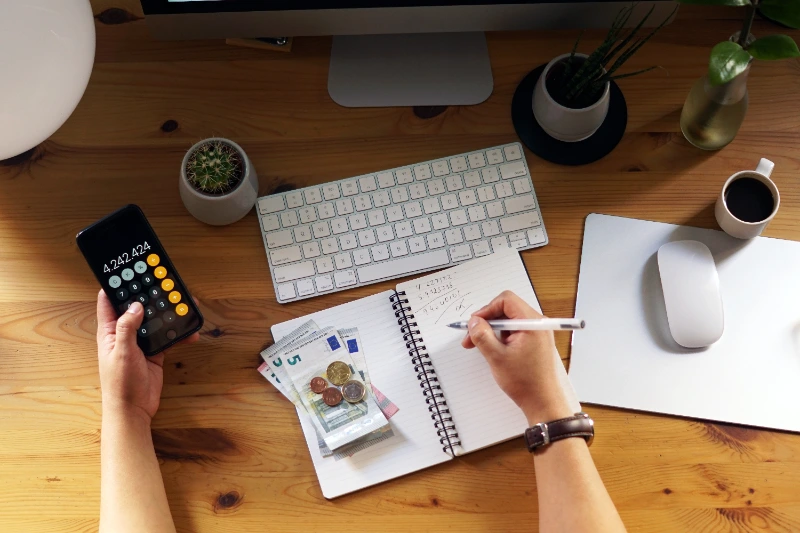 Vista superior de un emprendedor calculando finanzas personales con un teléfono inteligente y tomando notas en un cuaderno junto a billetes y monedas de euro sobre un escritorio de madera.