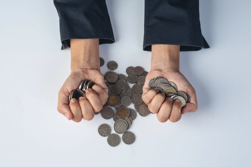 Vista cenital de una persona sosteniendo puñados de monedas sobre una superficie blanca junto a una pila dispersa, representando la acumulación de capital, el ahorro y la gestión financiera.