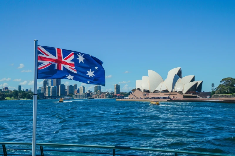 Bandera de Australia ondeando frente a la Bahía de Sídney con la Ópera de Sídney y el panorama urbano de fondo bajo un cielo despejado.
